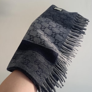 May include: A grey and charcoal scarf featuring a repeating interlocking GG logo pattern throughout the fabric. The scarf has a rectangular shape with a fringed edge on one end and a small rectangular Gucci brand label stitched near the top corner.