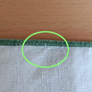 May include: Close-up of a white fabric with a green stitched border. The stitching is visible in the center of the image.