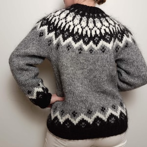 Puede incluir: Un suéter de lana gris y negro con un patrón tradicional escandinavo. El suéter tiene cuello redondo y mangas largas.