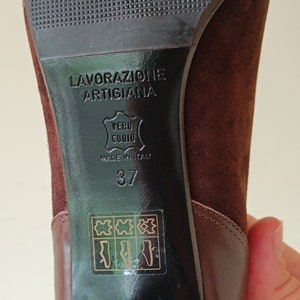 May include: Close-up of the bottom of a brown suede heeled shoe. The shoe has a black leather sole with the text "Lavorazione Artigiana" and "Vero Cuoio" in Italian. The shoe is size 37 and made in Italy.