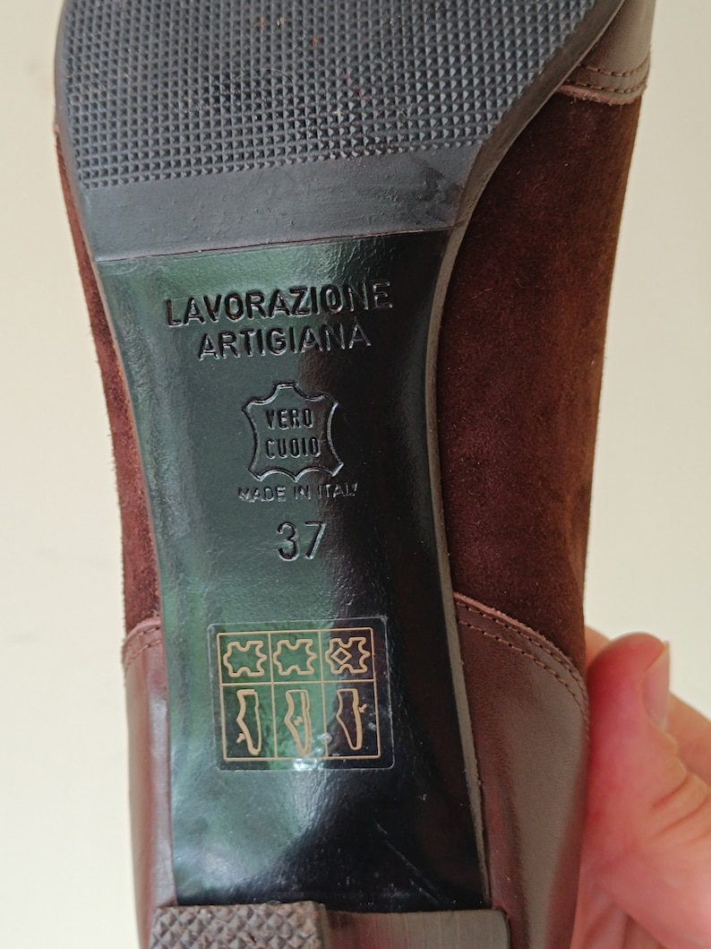 May include: Close-up of the bottom of a brown suede heeled shoe. The shoe has a black leather sole with the text "Lavorazione Artigiana" and "Vero Cuoio" in Italian. The shoe is size 37 and made in Italy.
