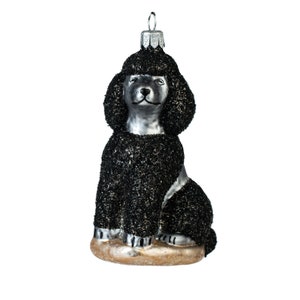 Peut inclure: Une décoration en verre en forme de caniche assis. Le caniche présente une finition texturée à paillettes noires sur les oreilles, la tête et le corps, avec un visage, un poitrail et des pattes argentés lisses. Il repose sur une base beige clair.