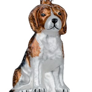 Könnte beinhalten: Ein Glasornament in Form eines sitzenden Beagle-Hundes mit braunen, schwarzen und weißen Abzeichen. Der Hund hat eine glänzende Oberfläche, eine schwarze Nase und eine silberne Metallkappe an der Oberseite mit einer kleinen Schlaufe zum Aufhängen. Die Ohren und der Rücken sind mit etwas weißem Glitzer bestäubt.