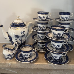 Può includere: Un servizio da tè in porcellana bianca con motivo willow blu e bordi dorati. Il set comprende una grande teiera, una lattiera e diverse tazze con piattini abbinati, decorati con illustrazioni blu raffiguranti alberi e uccelli.
