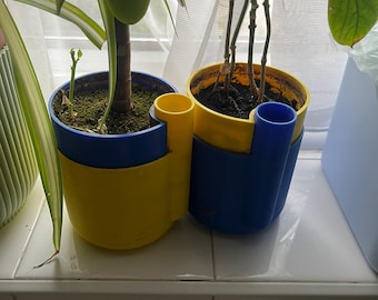Puede incluir: Dos plantas en macetas en macetas azules y amarillas. Las macetas tienen un diseño único con un asa incorporada. Una maceta es amarilla en la parte superior y azul en la inferior, y la otra es azul en la parte superior y amarilla en la inferior. Las plantas están en un alféizar.
