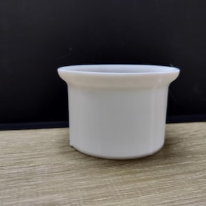 May include: A small, white ceramic container with a slightly flared rim. The cylindrical body has a smooth surface. The container sits on a wooden surface with a dark background. This item could be used for holding small items.