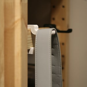 May include: A white plastic hook mounted on a wooden shelf with a gray leather strap hanging from it.