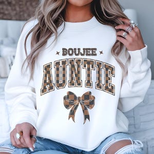 Może przedstawiać: Biała bluza z nadrukiem "BOOJEE AUNTIE" w czarno-brązową kratę. Pod tekstem znajduje się brązowo-czarna kokardka w kratę.