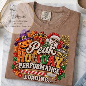Può includere: Una t-shirt marrone con una grafica raffigurante una zucca con un cappello da strega viola, un tacchino con occhiali da sole e Babbo Natale con occhiali da sole. Il testo recita Peak HOLIDAY PERFORMANCE 2027 LOADING con una barra di caricamento.