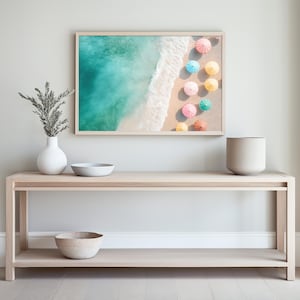 May include: A light wood console table with two shelves, a white ceramic vase with green branches, a white bowl, and a beige bowl. A framed print of a beach scene with colorful umbrellas hangs above the table.