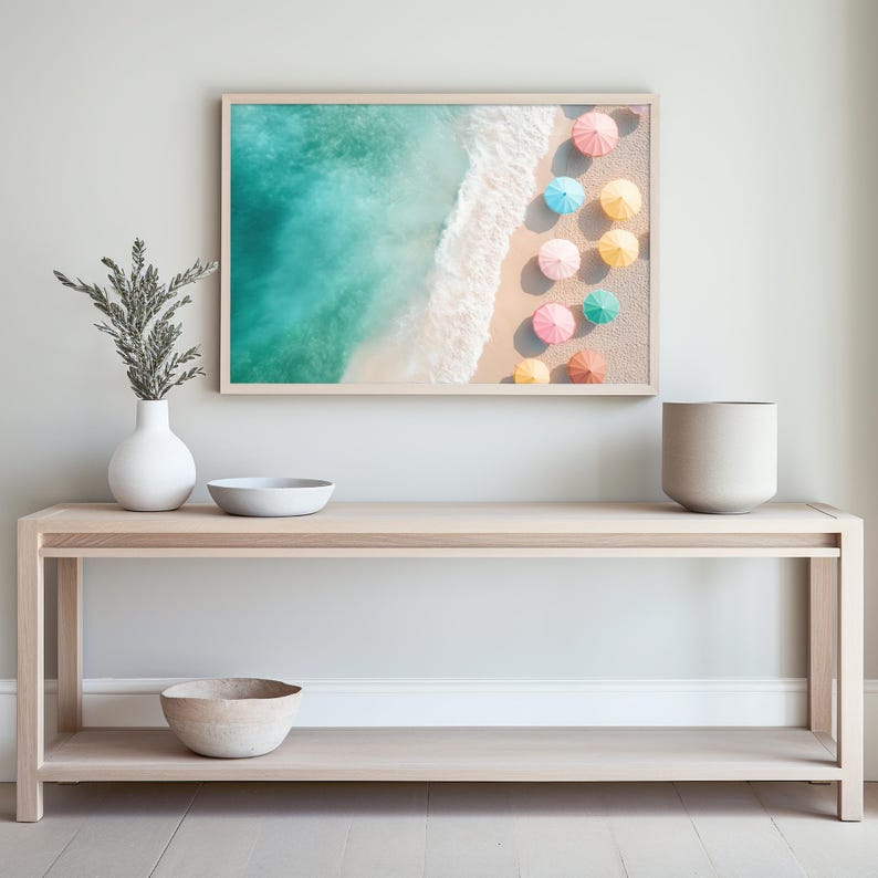 May include: A light wood console table with two shelves, a white ceramic vase with green branches, a white bowl, and a beige bowl. A framed print of a beach scene with colorful umbrellas hangs above the table.