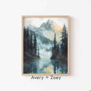 Puede incluir: Una pintura de acuarela de una escena de lago de montaña brumoso con altos árboles de hoja perenne que bordean la orilla. Las montañas están en el fondo, con un brillo dorado en la cima. La pintura está enmarcada en un marco de madera claro.