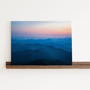 May include: A photograph of a mountain range at sunset. The mountains are layered and appear blue and purple in the fading light. The sky is a soft pink and orange.