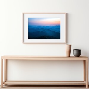 May include: A framed photograph of a mountain range with a blue and purple gradient sky at sunset. The photo is hanging on a white wall above a light wood console table with a vase and a bowl on top.