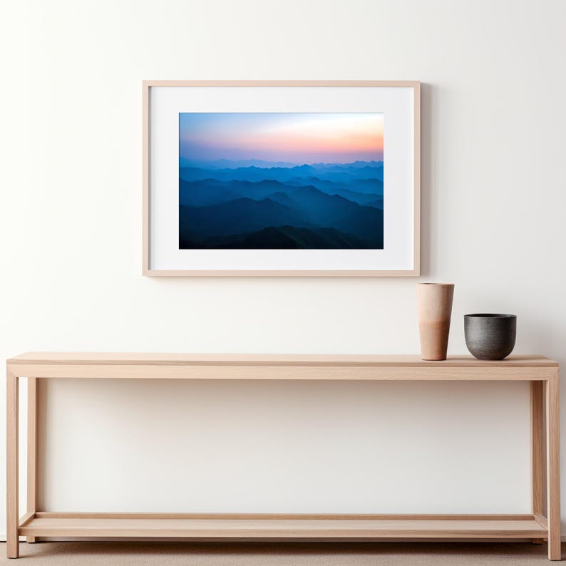 May include: A framed photograph of a mountain range with a blue and purple gradient sky at sunset. The photo is hanging on a white wall above a light wood console table with a vase and a bowl on top.