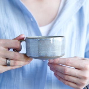 Op de afbeelding: Een keramische mok met een brede, cilindrische vorm en een kort handvat. De mok heeft een gespikkeld, lichtblauw en grijs glazuur met een gladde, glanzende afwerking aan de bovenzijde en een contrasterende, matte, donkergrijze basis aan de onderrand.