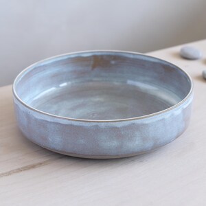 May include: A shallow, round ceramic bowl with a smooth, light blue and grey glazed finish. The bowl features a wide, flat base and slightly curved walls, showcasing a subtle variation in color tones across the interior and exterior surfaces.