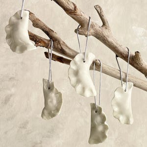 May include: Five white ceramic pierogi ornaments hanging from a natural wooden branch. Each ornament has a unique shape and is suspended by a silver wire. The ornaments are a creamy white colour.