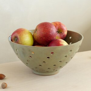 Può includere: Una fruttiera in ceramica verde piena di mele rosse e gialle. La ciotola ha fori distanziati uniformemente. Diverse nocciole sono sparse intorno alla ciotola su una superficie chiara.