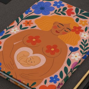 Op de afbeelding: Een dagboek met een bloemmotief en een illustratie van een zwangere persoon. De omslag heeft een perzikkleurige achtergrond met blauwe, oranje en witte bloemen. De illustratie toont een persoon met een baby in haar buik. Gouden en zwarte potloden liggen naast het dagboek.