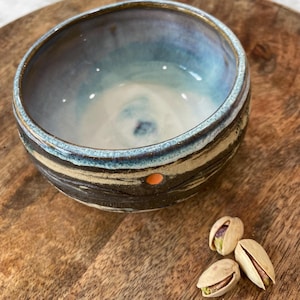 May include: A handmade ceramic bowl with a blue and white interior and a dark exterior with a textured design. The bowl has a small orange dot on the side and is next to three open pistachio nuts.