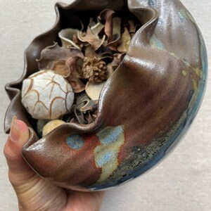 May include: A handcrafted ceramic bowl with a ruffled edge, filled with dried botanicals and decorative stones. The bowl features a glossy, mottled glaze in shades of brown, red, and blue, creating a unique, organic aesthetic.