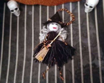 Puede incluir: Una pequeña muñeca de bruja hecha de hilo con un sombrero negro, un vestido negro y una escoba. La muñeca tiene una cara blanca con labios rojos pintados y los ojos cerrados. La muñeca está sentada sobre un fondo de tela a rayas.