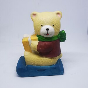 Puede incluir: Una figurita de cerámica de un oso de peluche amarillo que lleva un suéter rojo y una bufanda verde, sosteniendo una caja de regalo amarilla y sentado en un trineo azul.