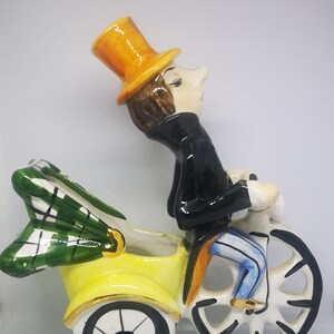 May include: A ceramic figurine of a man in a yellow top hat riding a yellow tricycle with a green plaid seat. The man is wearing a black jacket and blue pants.