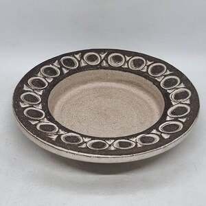 Può includere: Un piccolo piatto rotondo in ceramica con una glassa bianca e un motivo geometrico marrone attorno al bordo.