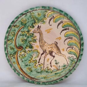 Könnte beinhalten: Dekorative Keramikplatte mit handgemaltem Design. Die Platte zeigt einen laufenden Hirsch in einer naturalistischen Umgebung mit grünem Laub und einem hellrosa Himmel. Der Rand ist gewellt und grün und braun bemalt.