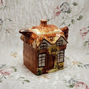 Peut inclure: Une théière en céramique brune et blanche en forme de cottage avec un toit de chaume. La théière a un bec verseur et une poignée. Le cottage a des fenêtres et une porte.
