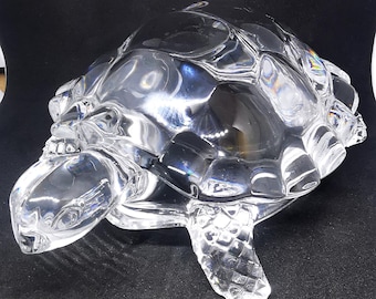May include: A clear glass turtle figurine with detailed shell and flipper features. The sculpture is transparent, allowing light to pass through, and sits on a dark surface. The turtle is a decorative item.