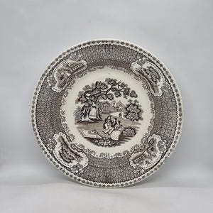 Peut inclure: Une assiette en céramique blanche avec un motif brun et gris. Le motif représente une scène avec des personnes et des arbres. L'assiette a un bord festonné.
