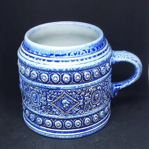 Może przedstawiać: Niebiesko-biały ceramiczny kufel do piwa ze skomplikowanymi wzorami reliefowymi. Cylindryczny kubek posiada uchwyt i szczegółowe wzory postaci, kształtów geometrycznych i motywów kwiatowych. Wnętrze kufla jest białe.