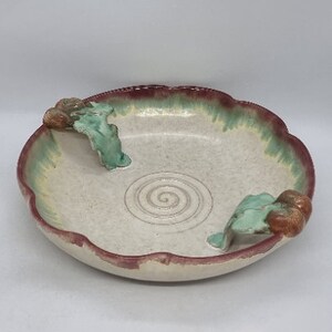 Pode incluir: Uma tigela de cerâmica branca com alças em forma de folhas verdes e marrons. A tigela tem uma borda ondulada e um desenho em espiral no centro. A tigela é envidraçada em branco, verde e marrom.