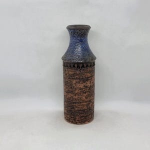 Peut inclure: Un vase en céramique texturé et de grande taille, avec un corps cylindrique et un col évasé. Le vase présente une base marron, un col bleu-gris et une bande décorative près du sommet. Le vase a un aspect rustique et artisanal.
