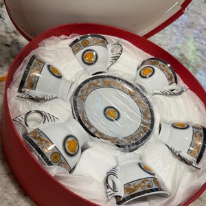 May include: A set of six white porcelain teacups and saucers with gold and black decorative accents, presented in a red round gift box. Each cup features a gold emblem and ornate detailing. The set includes a central saucer.