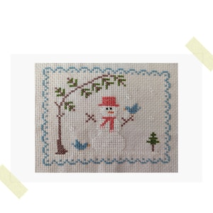 Op de afbeelding: Borduurwerk met kruissteek van een sneeuwpop met een rode muts en sjaal, twee blauwe vogels, een groene boom en een bruine boom met groene bladeren. Het ontwerp is omgeven door een blauwe en witte rand.