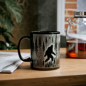 May include: Black coffee mug featuring a silhouette of Bigfoot walking through a forest of pine trees. The mug has a black handle and rim. Steam rises from the mug, suggesting a hot beverage. The mug sits on a wooden surface.