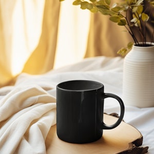 May include: A black ceramic mug sits on a wooden slice, resting on a white bedspread. A vase with green leafy branches is in the background.