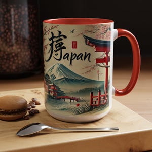 May include: A white ceramic mug with a red rim and handle. The mug features a Japanese-inspired design with a mountain, cherry blossoms, a red gate, and the word "Japan" in English and Japanese characters.