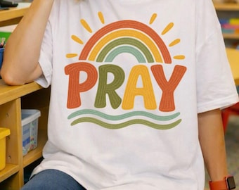 May include: A white short-sleeved t-shirt featuring a graphic print of a stylized rainbow in shades of orange, yellow, and green. Below the rainbow, the word PRAY is printed in large, bold, textured letters, with two wavy lines underneath.
