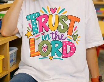 May include: A white short-sleeved t-shirt featuring a colorful graphic print with the text "TRUST in the LORD" in bold, multi-colored letters. The design includes various patterns like polka dots and stripes, accented by small hearts and stars.