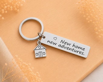 Op de afbeelding: Een zilverkleurige metalen sleutelhanger met een ronde ring, een kleine bedel in de vorm van een huis en een rechthoekige hanger gegraveerd met een hartsymbool en de tekst New home new adventures. Het ontwerp is strak en metaalachtig.