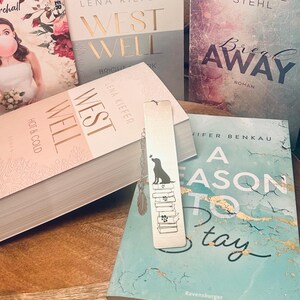 Könnte beinhalten: Nahaufnahme eines Stapels Bücher mit einem goldenen Lesezeichen, das eine schwarze Hundesilhouette und einen Stapel Bücher zeigt. Das Lesezeichen liegt auf einem Buch mit dem Titel "A Season to Stay" von Jennifer Benkau. Die anderen Bücher tragen die Titel "West Well" von Lena Kiefer und "Break Away" von Anabelle Stehl.