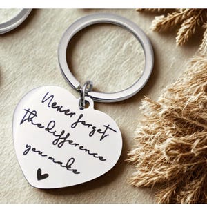 Könnte beinhalten: Ein herzförmiger silberner Schlüsselanhänger mit der Aufschrift "Never forget the difference you make" in schwarzer Schrift. Unter dem Text befindet sich ein kleines schwarzes Herz. Der Schlüsselanhänger hat einen silbernen Ring.
