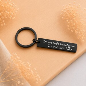 Könnte beinhalten: Ein schwarzer Metallschlüsselanhänger mit einem rechteckigen Anhänger, der mit dem weißen Text "Drive safe handsome. I love you." und einem kleinen Herzsymbol graviert ist. Der Schlüsselanhänger besteht aus einem Ring und einem flachen Anhänger.