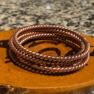 May include: Three copper wire bracelets, each with a twisted design, stacked on top of each other on a wooden surface.