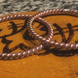May include: Two copper wire bracelets with a braided design. The bracelets are on a wooden surface with black markings.